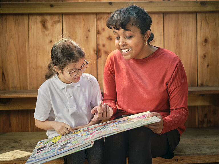 a woman in a red top reads a picture book with a young girl in a white top. They are pointing at a picture together.