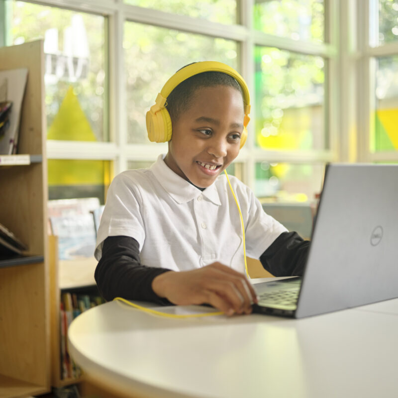 Boy in an online reading session. He's wearing yellow headphones and is smiling at the laptop.