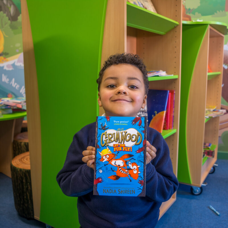 Boy in blue school uniform, smiling and holding up a book.