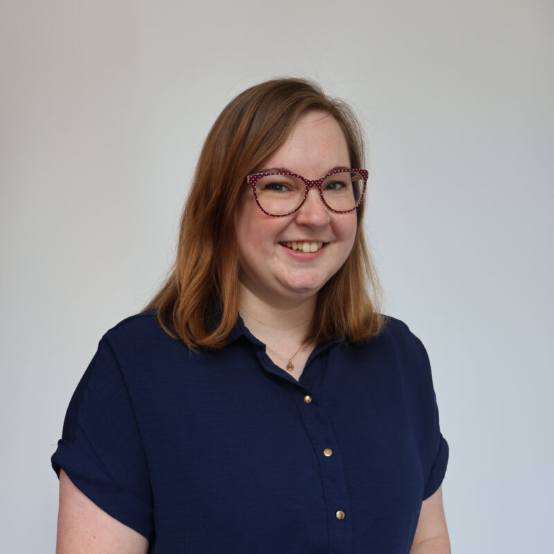 She has short brown hair and glasses, she's wearing a blue blouse and is smiling at the camera