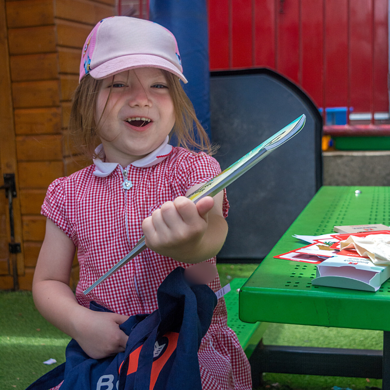 Girl in red checked dress wearing a pink hat holding a book from Bookmark's Story Starter Programme pack.