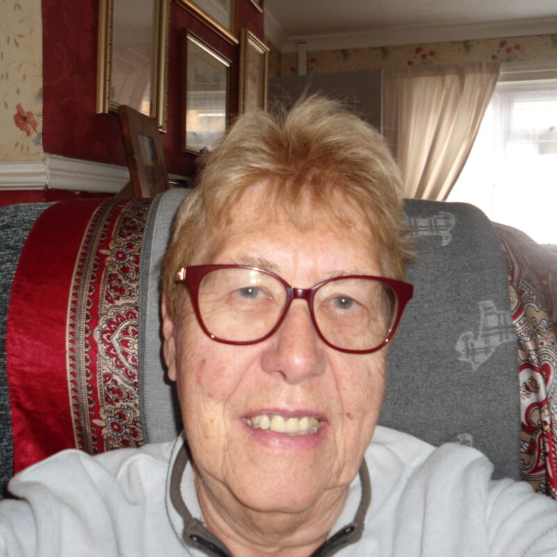 Selfie shot of older female reading volunteer in a chair, smiling at the camera. She has short light brown hair and is wearing glasses.