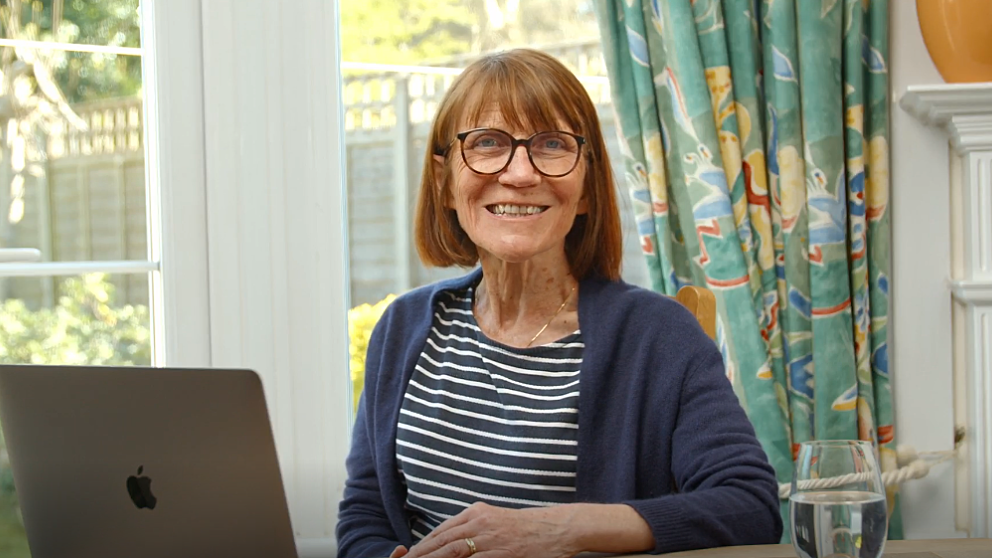 A Bookmark volunteer sits at a table with a laptop in front of them, wearing a striped shirt and a cardigan. A glass of water is on the table, and behind them are patterned curtains and a bright window looking out onto a garden.