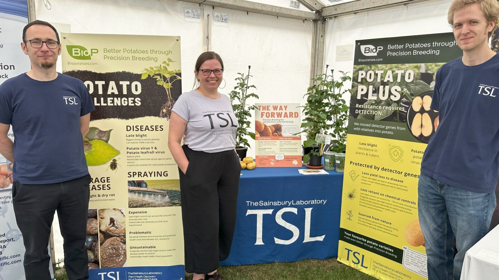 three scientists standing in front of the biopoatoes stand and banners within a white marquee