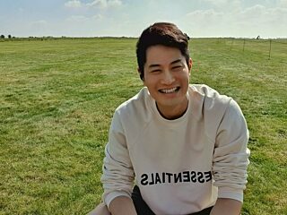 Photo of Bruno Ngou crouching down in a grass field. Bruno is wearing a white sweatshirt with the word ESSENTIALS printed in mirror-image across the front.