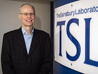 Photograph of Dr Stephen Bornemann standing in front of a sign at the entrance to The Sainsbury Laboratory