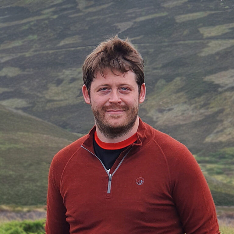Photo of George Deeks wearing a red sweatshirt against a background of hills